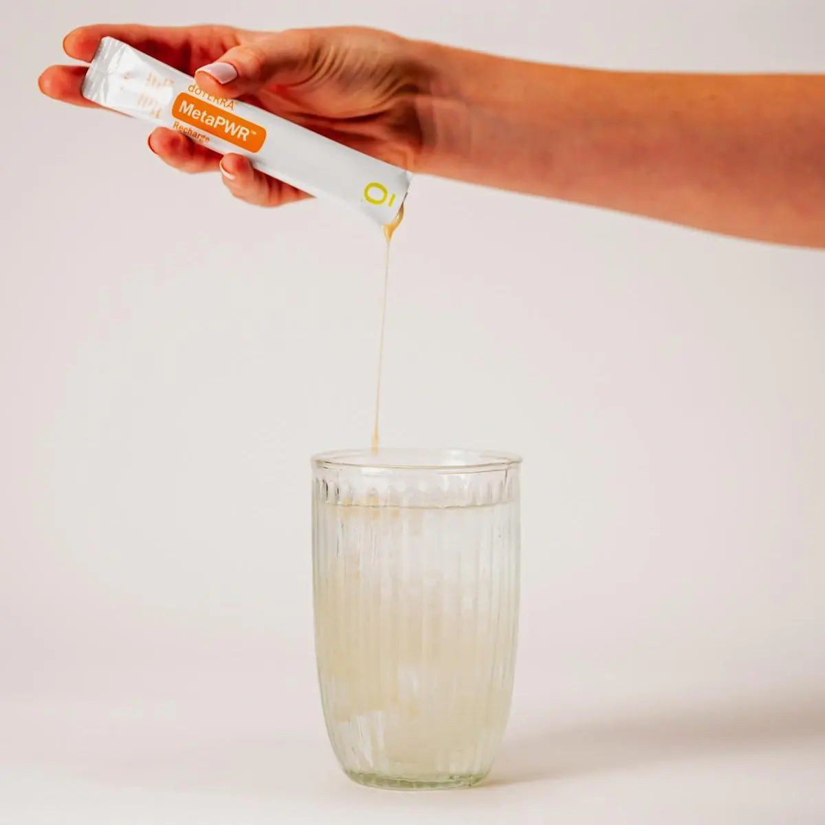 Mano virtiendo el líquido dorado de un sobre de dōTERRA MetaPWR Recharge Lemon-Lime en un vaso de agua con hielo.