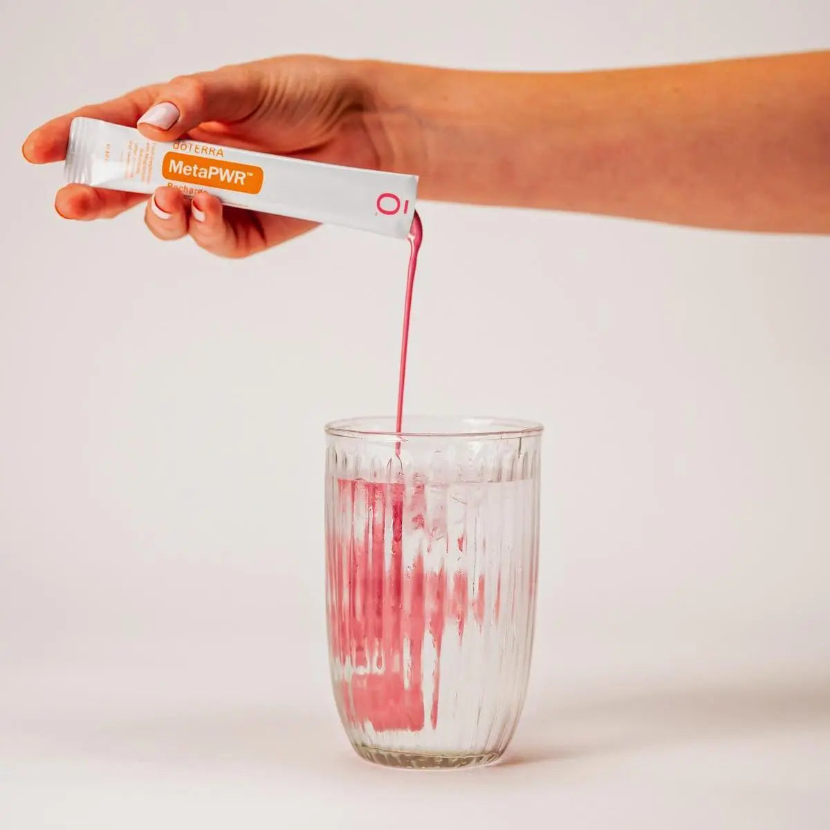 Mano virtiendo el líquido rosa de un sobre de dōTERRA MetaPWR Recharge Wild Strawberry en un vaso de agua con hielo.
