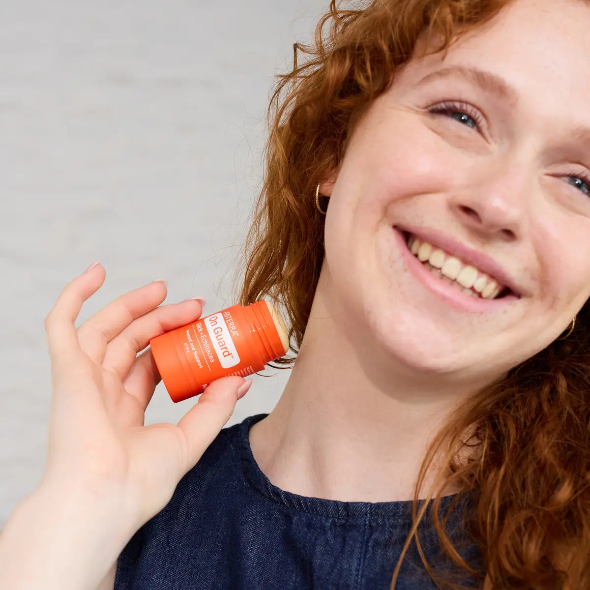 Mujer pelirroja sonriente sosteniendo el dōTERRA On Guard Stick con Equinácea junto al cuello.