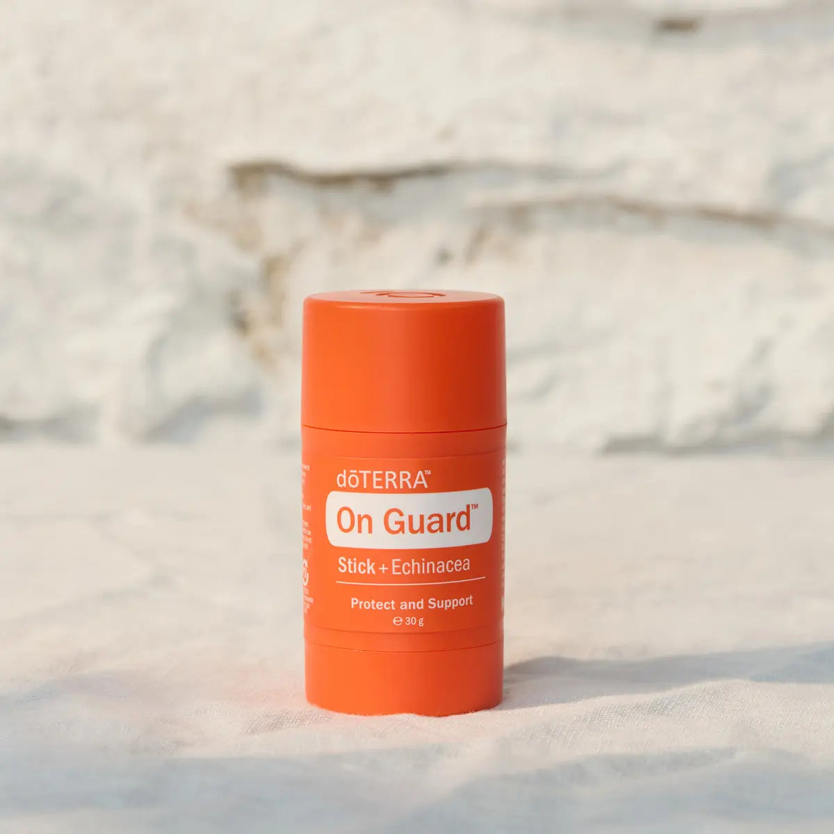 Envase en barra dōTERRA On Guard Stick con Equinácea en pie sobre tejido con fondo de pared de piedra clara.