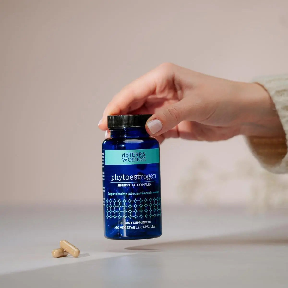 Mano sosteniendo frasco azul de dōTERRA Phytoestrogen Essential Complex con dos cápsulas sobre la mesa.