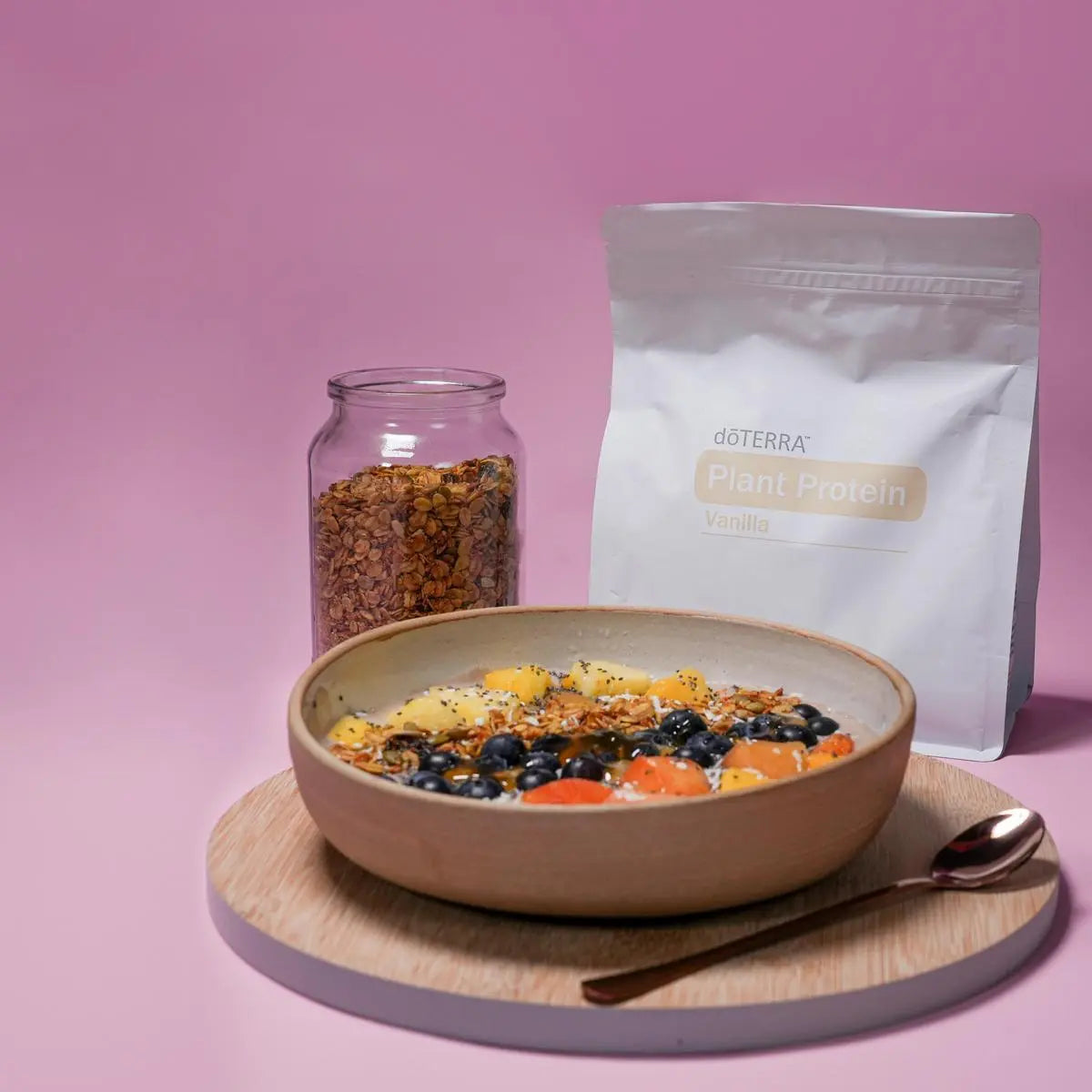 Bolsa de dōTERRA Plant Protein Vanilla junto a un bowl de desayuno con fruta, arándanos y granola sobre fondo rosa.