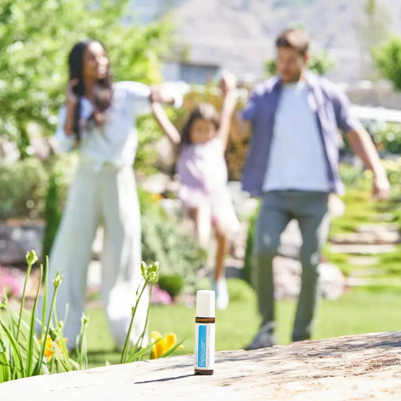 Frasco dōTERRA Rescuer Touch 10ml en primer plano con familia jugando al aire libre en el jardín desenfocada al fondo.