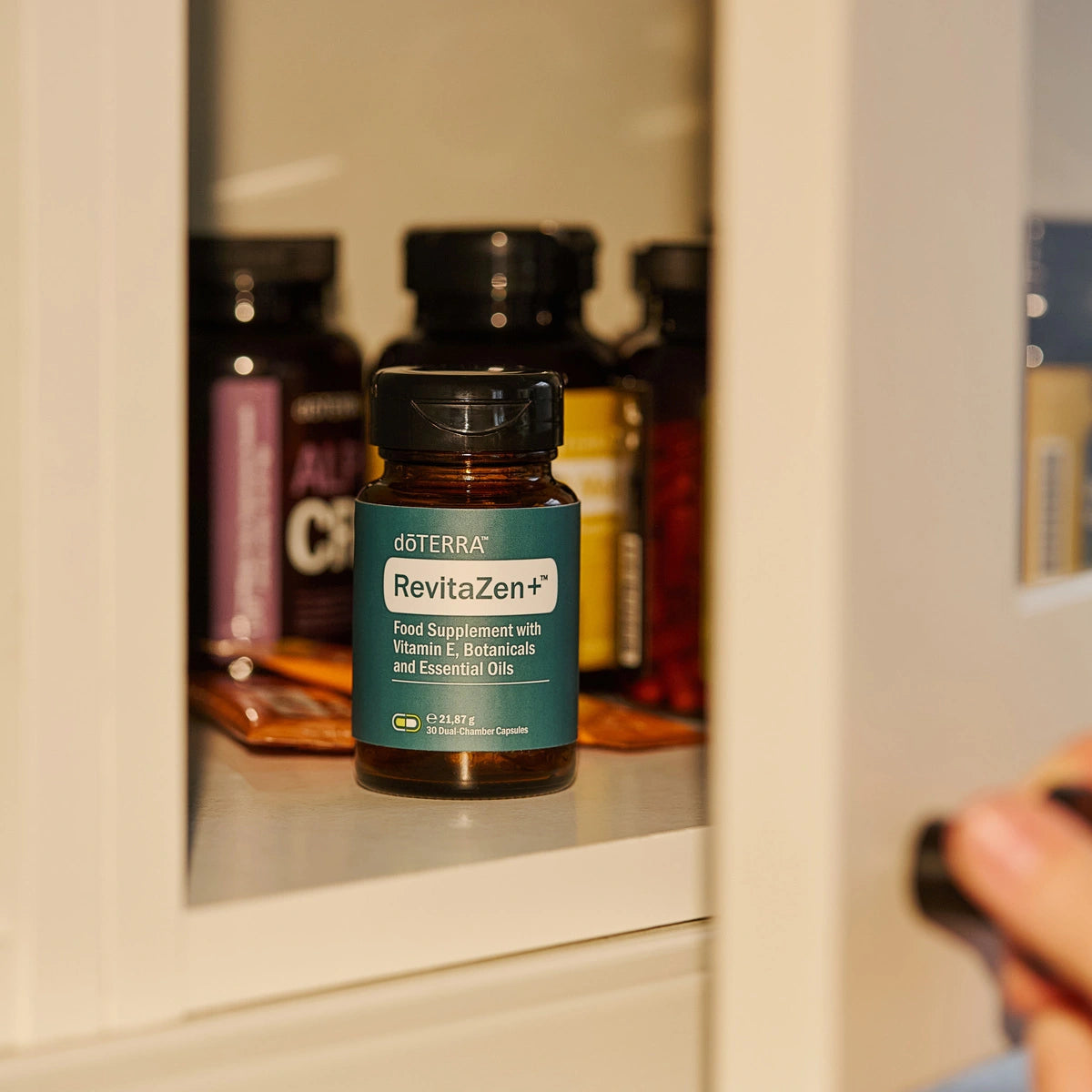 Frasco de dōTERRA RevitaZen Plus en un armario de suplementos blanco, con mano abriendo la puerta.