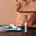 Frasco roll-on dōTERRA Tamer Touch 10ml sobre mesa ao lado de caderno, lápis de madeira e mochila de pele ao fundo.
