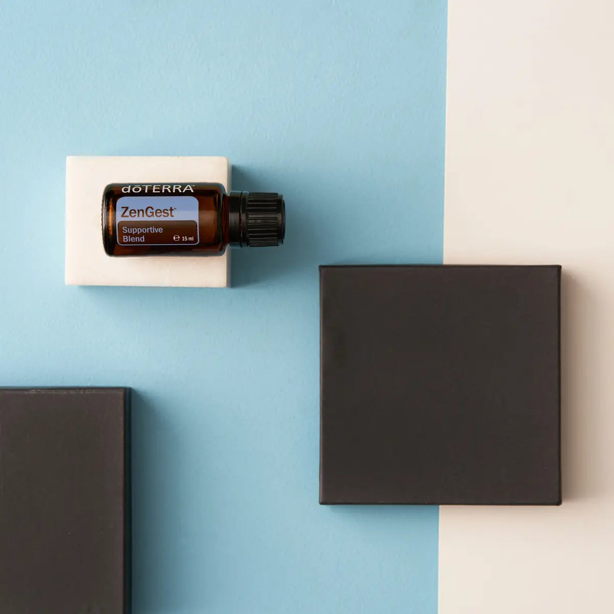 Frasco de aceite esencial dōTERRA ZenGest 15ml en horizontal sobre soporte de mármol blanco en fondo azul y beige geométrico.