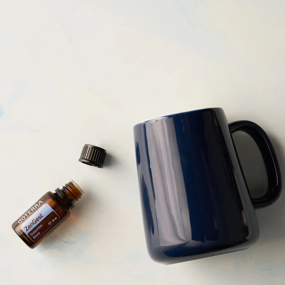 Vista superior del frasco de aceite esencial dōTERRA ZenGest 15ml abierto junto a su tapón y una taza azul oscura.