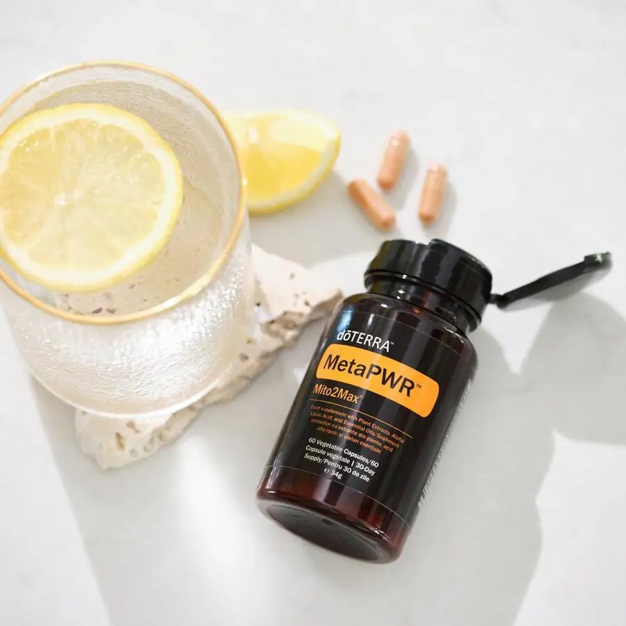 MetaPWR Mito2Max doTERRA en plano cenital sobre marmol blanco con vaso de agua con rodaja de limon y capsulas beige sobre base de piedra