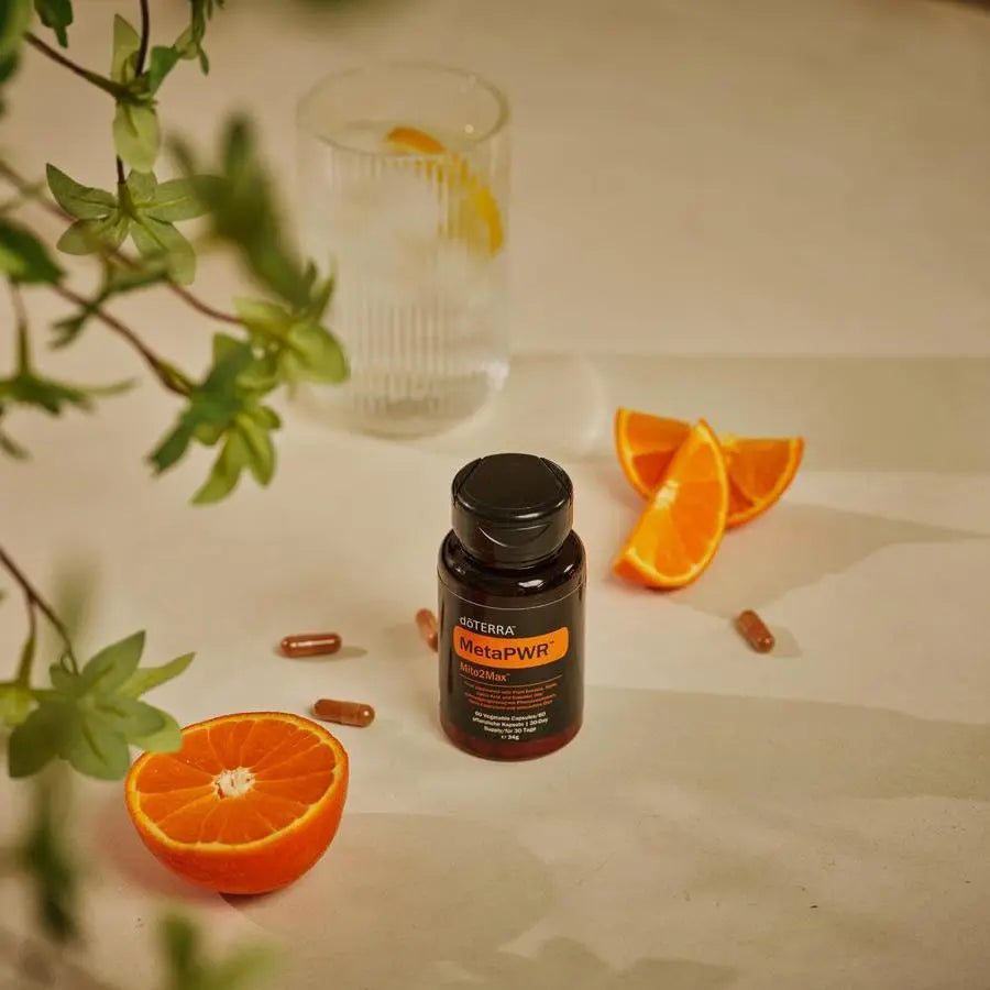 MetaPWR Mito2Max doTERRA sobre superficie beige con gajos de naranja, vaso de agua con limon, ramas de planta verde y capsulas sueltas