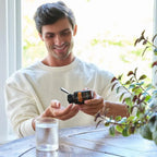 Hombre con jersey blanco sonriendo mientras vierte capsulas de MetaPWR Mito2Max doTERRA en la mano, con vaso de agua sobre mesa rustica