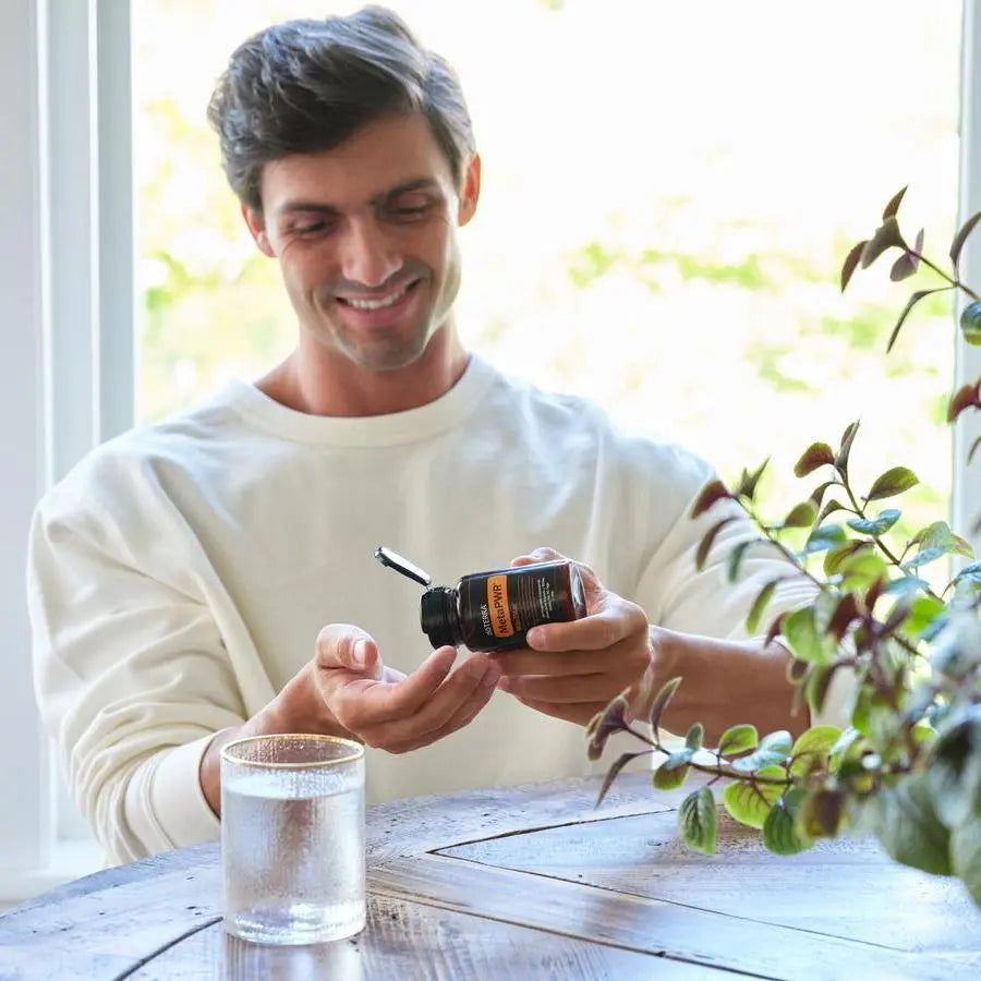 Hombre con jersey blanco sonriendo mientras vierte capsulas de MetaPWR Mito2Max doTERRA en la mano, con vaso de agua sobre mesa rustica