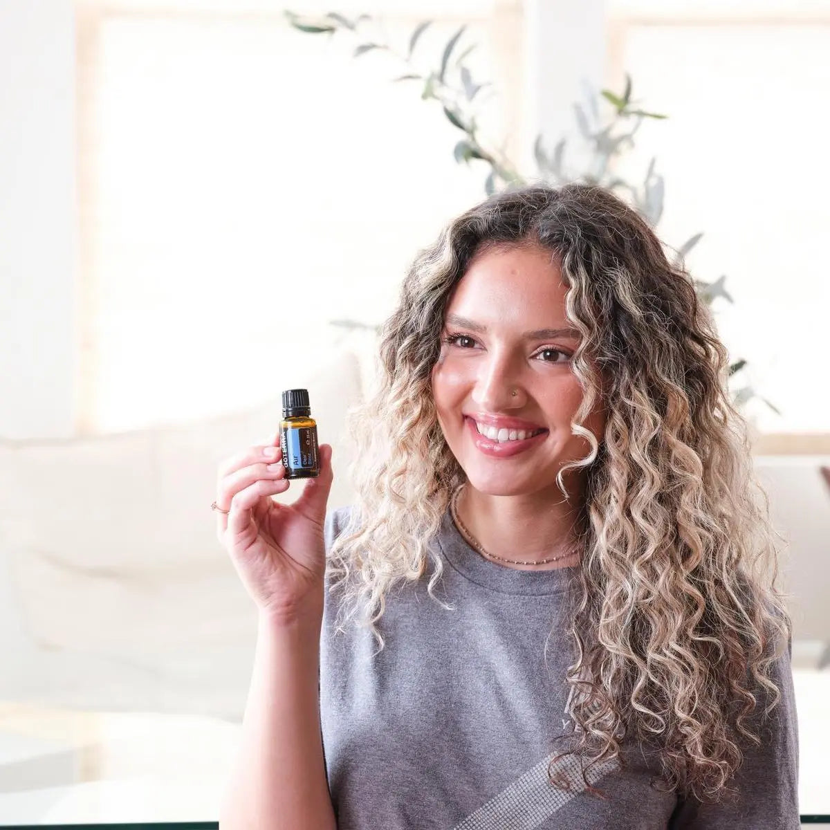Mujer rubia de pelo rizado sosteniendo el frasco de Mezcla Air dōTERRA 15ml en salón blanco con plantas