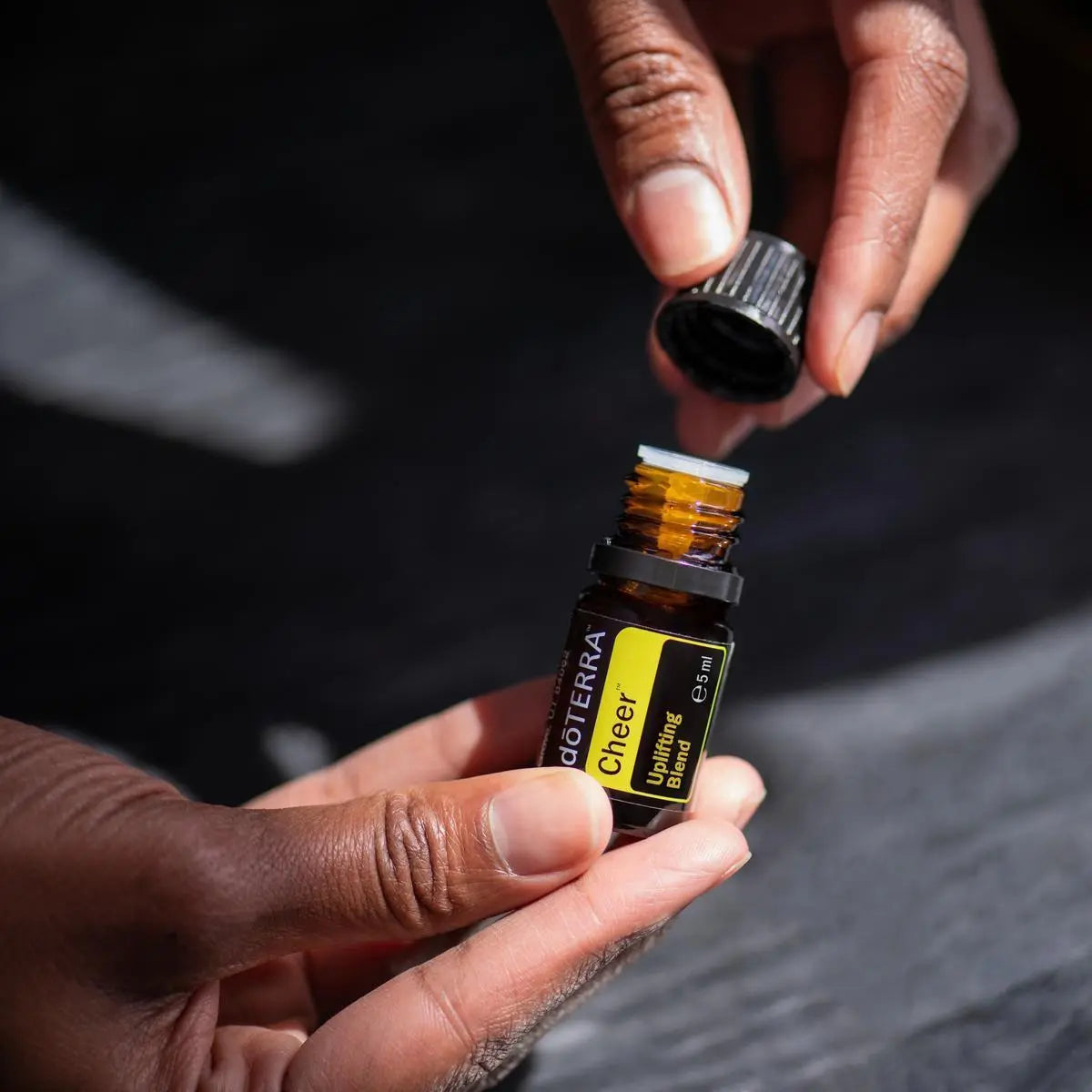 Mezcla dōTERRA Cheer 5ml — dos manos abriendo el frasco sobre fondo oscuro dramático