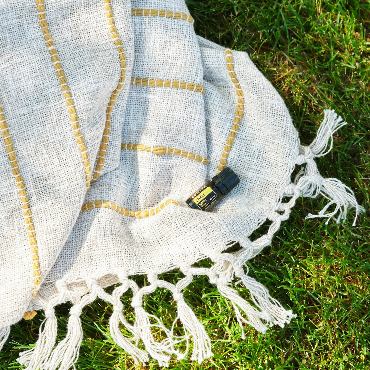 Mezcla dōTERRA Cheer 5ml — frasco sobre manta de lino beige con flecos dorados y hierba verde