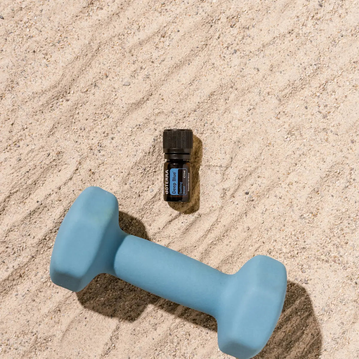 Mezcla dōTERRA Deep Blue 5ml — frasco sobre arena junto a mancuerna azul con luz solar, contexto deportivo, vista cenital
