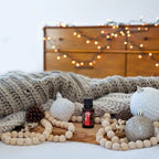Mezcla dōTERRA Holiday Joy 15ml — frasco sobre manta gris con bolas de navidad plateadas, piñas y luces en bokeh