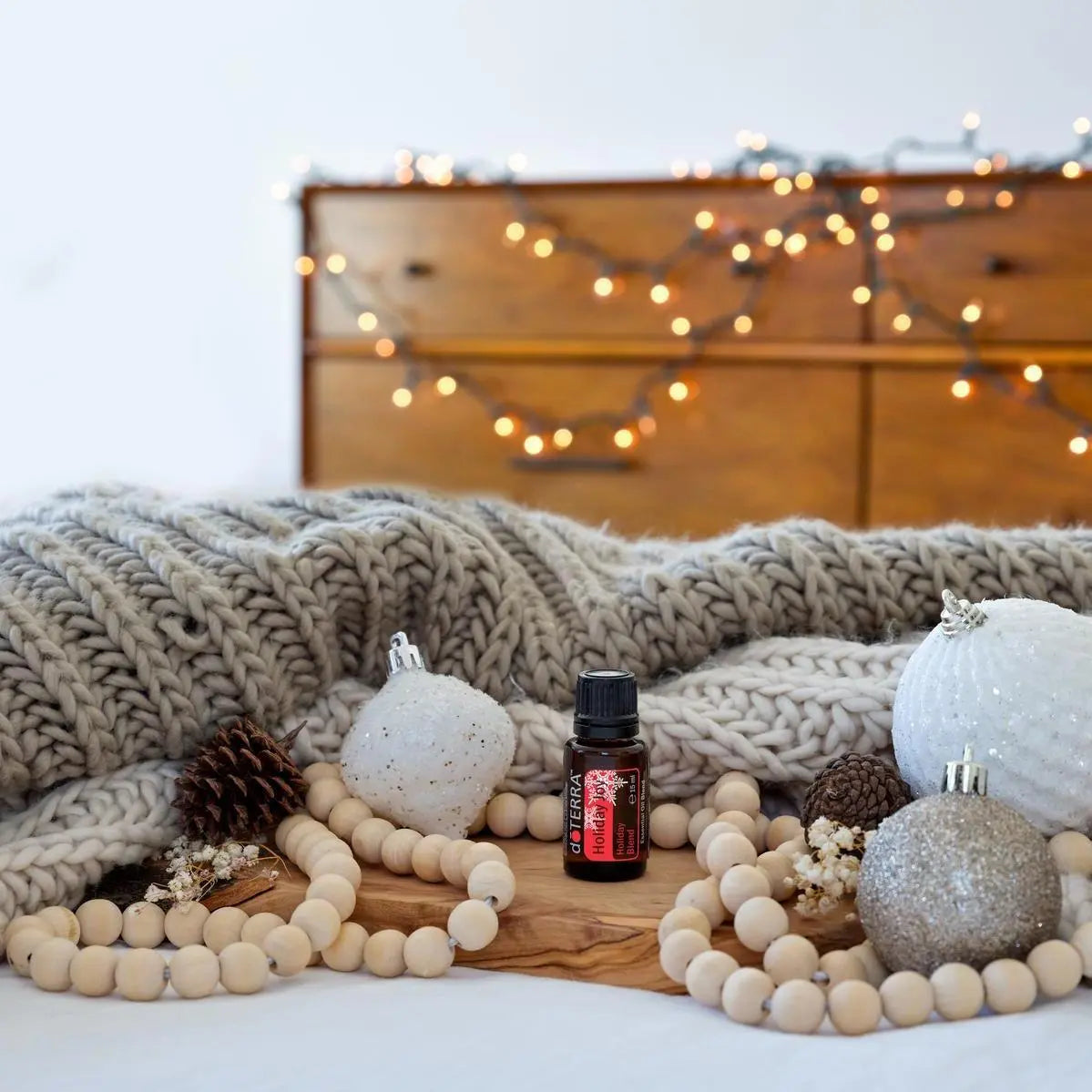 Mezcla dōTERRA Holiday Joy 15ml — frasco sobre manta gris con bolas de navidad plateadas, piñas y luces en bokeh