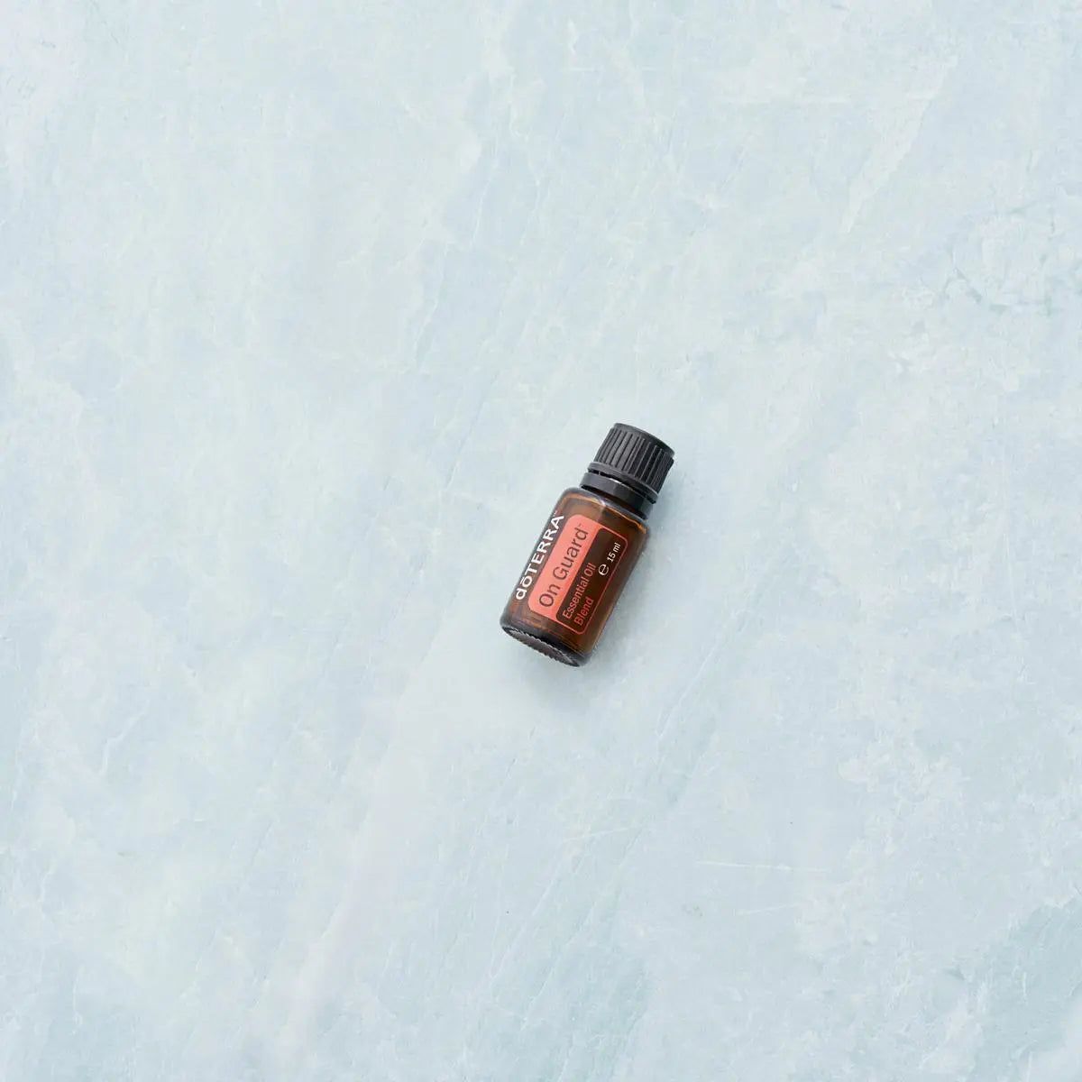 Mezcla dōTERRA On Guard 15ml — frasco tumbado sobre mármol azul claro, vista cenital minimalista