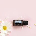 Mezcla dōTERRA Purify 15ml — frasco tumbado entre pétalos de margarita blanca sobre fondo rosa, vista cenital
