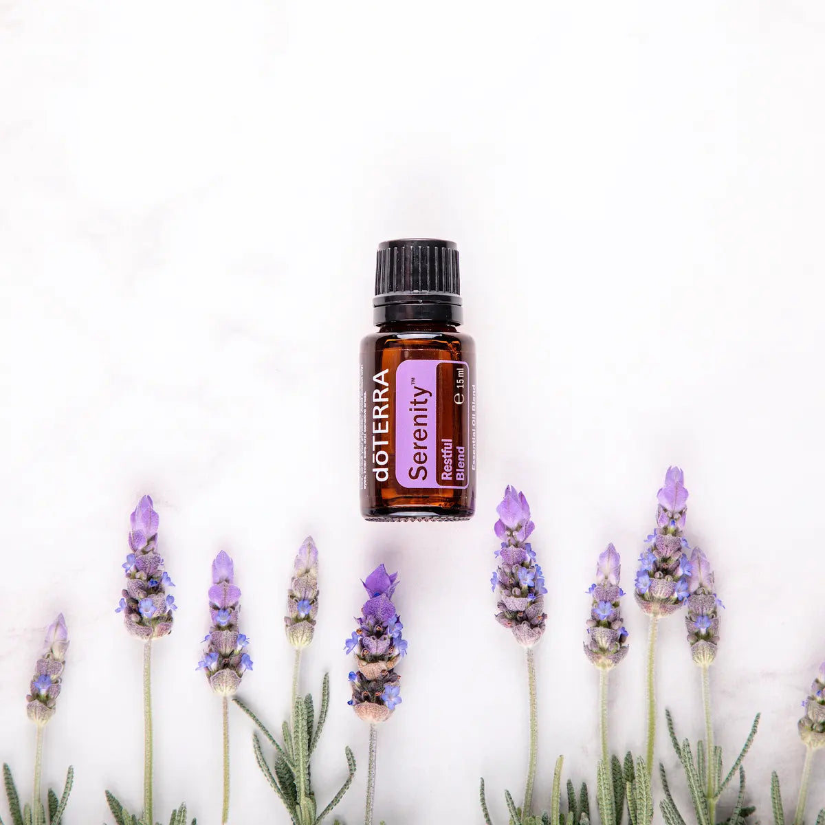 Mezcla dōTERRA Serenity 15ml — frasco flotando en fila entre tallos de lavanda sobre mármol blanco, vista cenital