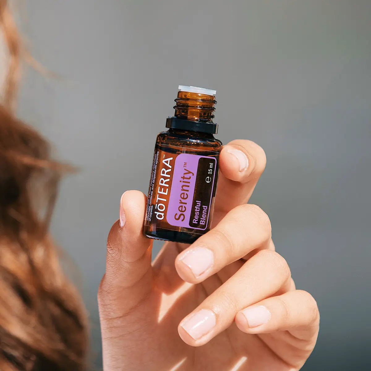 Mezcla dōTERRA Serenity 15ml — mano pelirroja sosteniendo el frasco abierto en primer plano con fondo gris