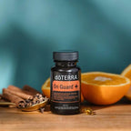 On Guard+ Softgels doTERRA sobre superficie de madera calida junto a mitades de naranja, ramas de canela y clavos de especia en cuchara dorada, fondo azul verdoso