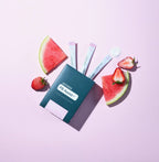 PB Assist+ doTERRA sobres individuales sobre fondo rosa pastel rodeados de sandia y fresas frescas cortadas en composicion de estudio