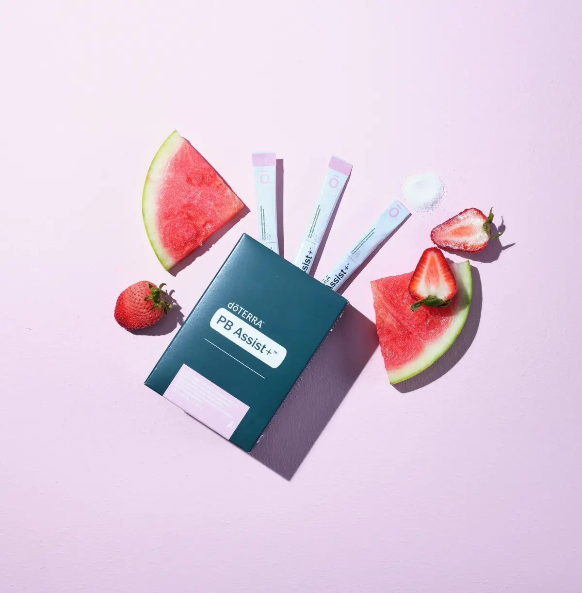 PB Assist+ doTERRA sobres individuales sobre fondo rosa pastel rodeados de sandia y fresas frescas cortadas en composicion de estudio