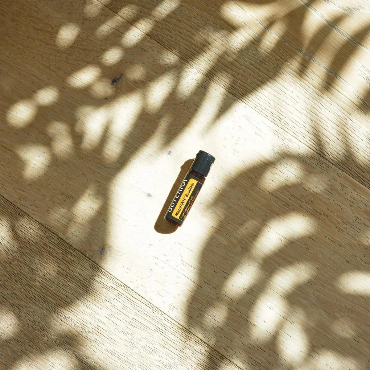 Perlas MetaPWR dōTERRA — frasco en suelo de parque de madera con sombras de árbol y luz solar, vista cenital