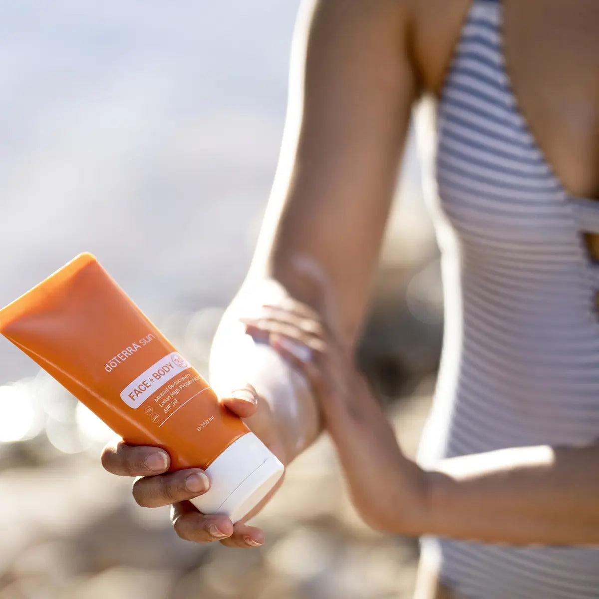 Protector solar Sun Face+Body dōTERRA SPF 30 — tubo con mujer aplicando en el brazo en la playa