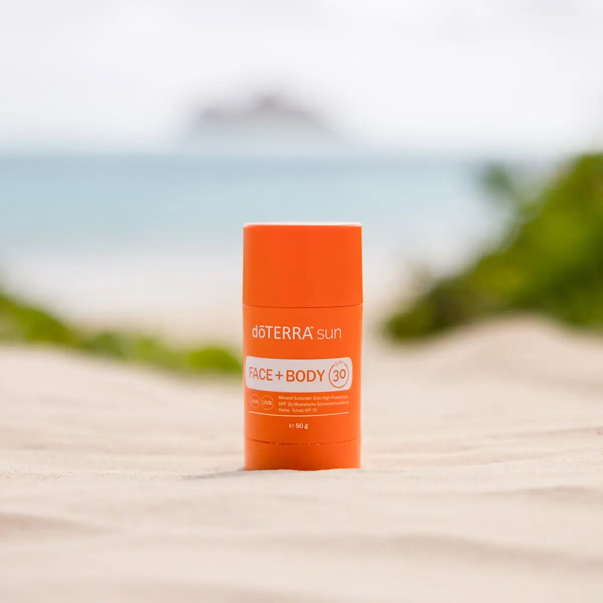 Protector solar Sun Face+Body Stick dōTERRA SPF 30 50g — en arena de playa con mar e isla al fondo