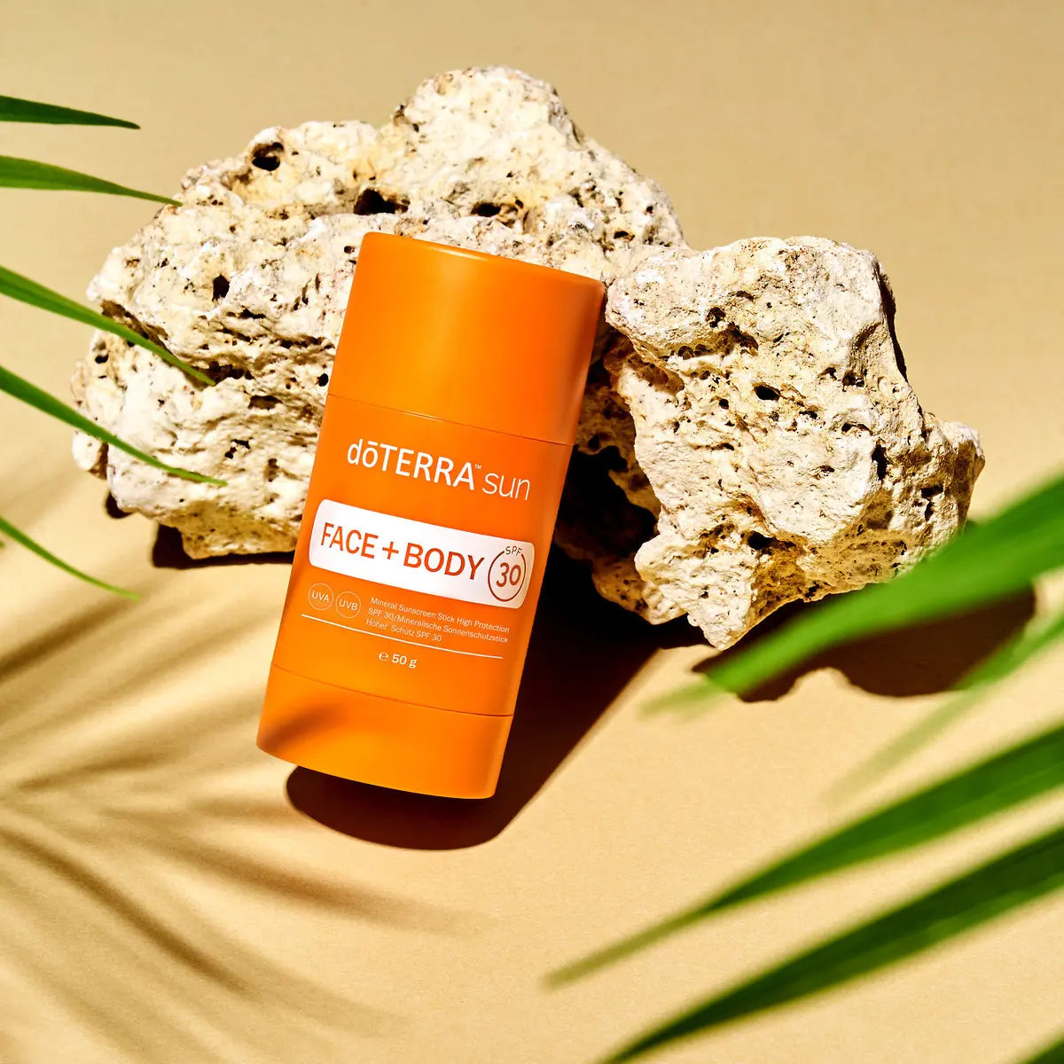 Protector solar Sun Face+Body Stick dōTERRA SPF 30 50g — sobre rocas de coral con hojas de palmera y arena