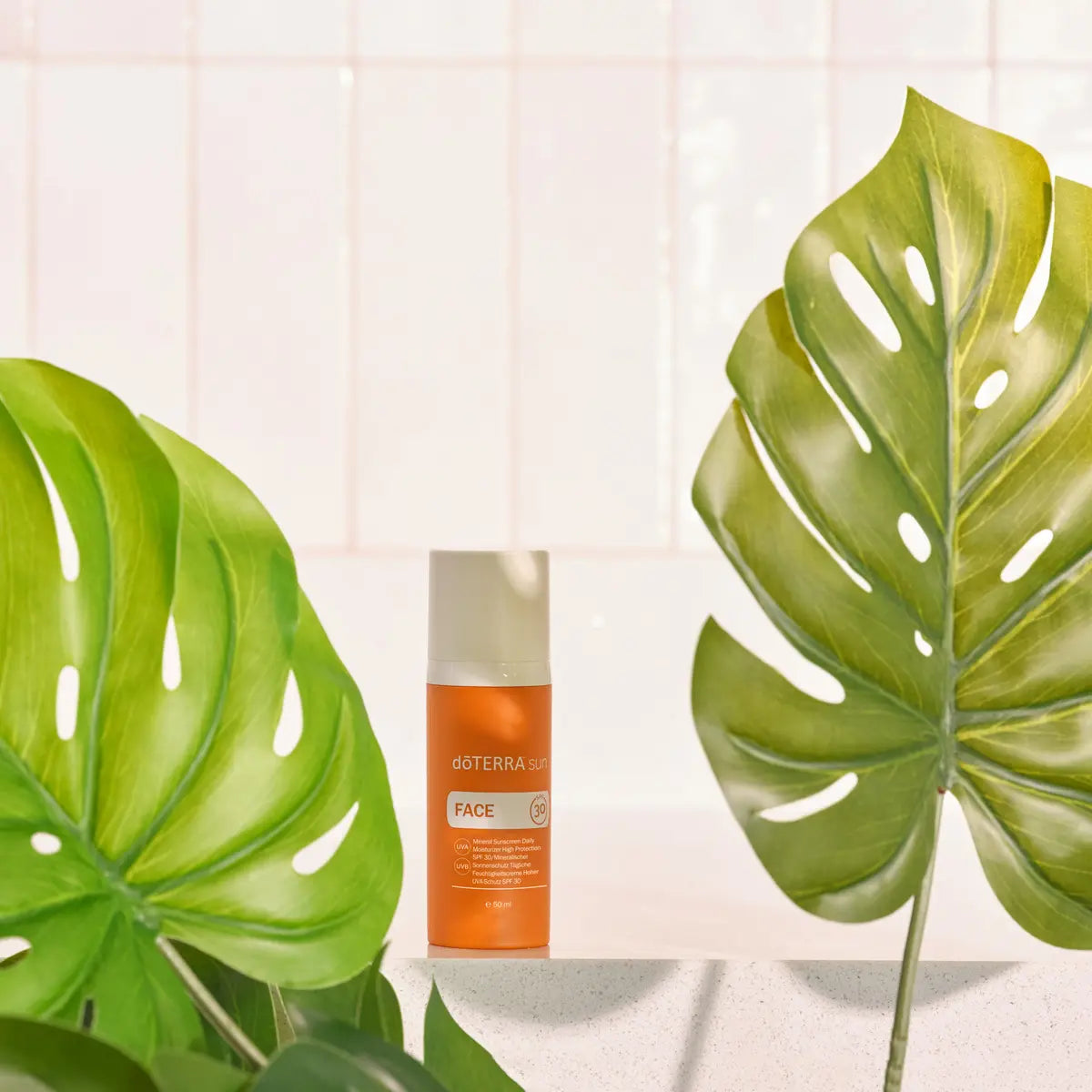 Protector solar Sun Face dōTERRA SPF 30 — frasco sobre hojas de monstera y azulejo blanco