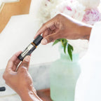 Manos de piel oscura sujetando Copaiba Touch dōTERRA 10ml con peonías rosadas de fondo