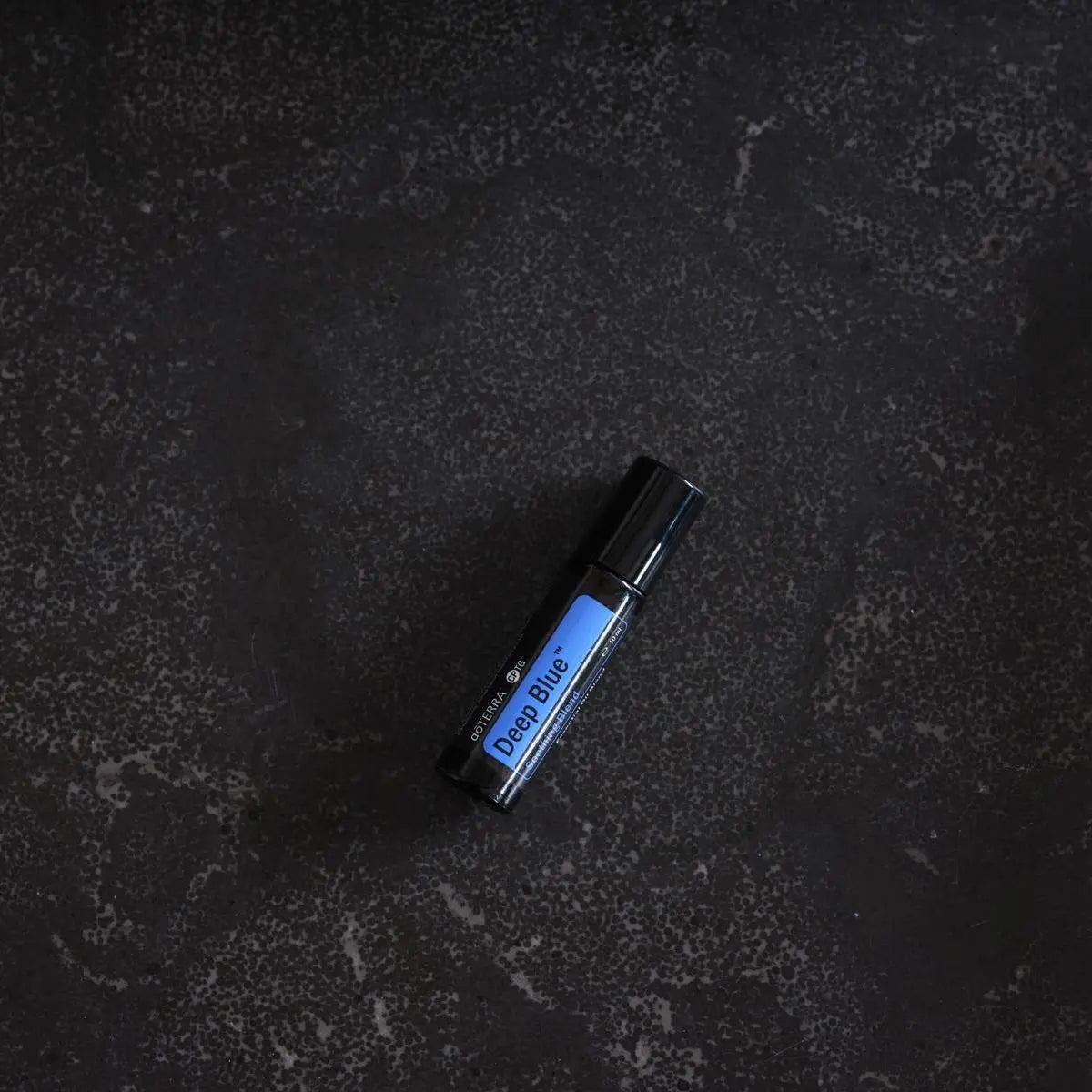 Roll-on dōTERRA Deep Blue 10ml — tumbado sobre granito negro texturizado, vista cenital