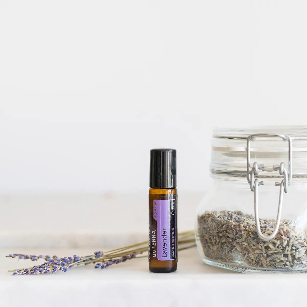 Lavanda Touch dōTERRA roll-on 10ml junto a lavanda seca en tarro de cristal con ramita sobre fondo blanco