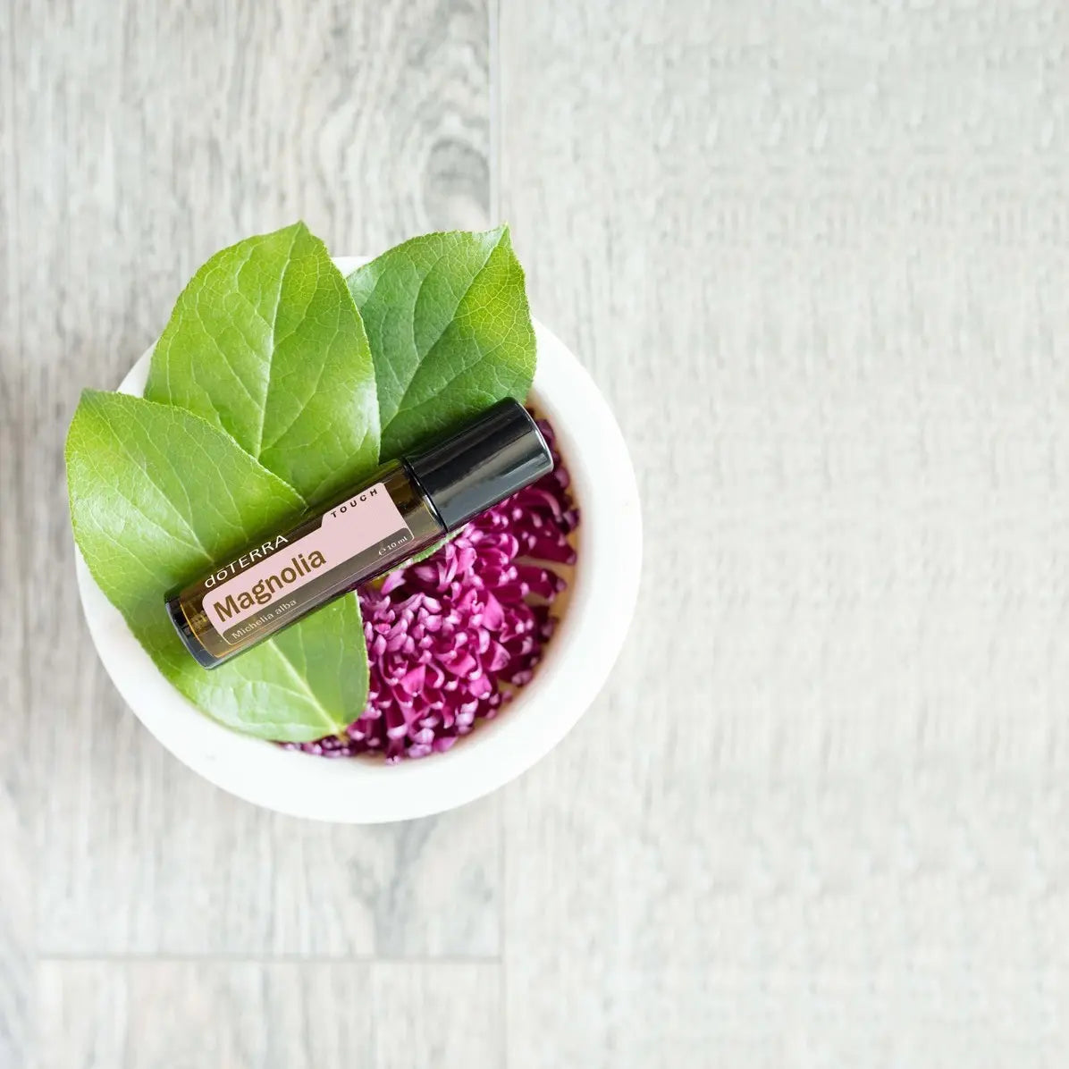 Roll-on dōTERRA Magnolia Touch 10ml — en cuenco blanco rodeado de flores lila y hojas verdes, vista cenital