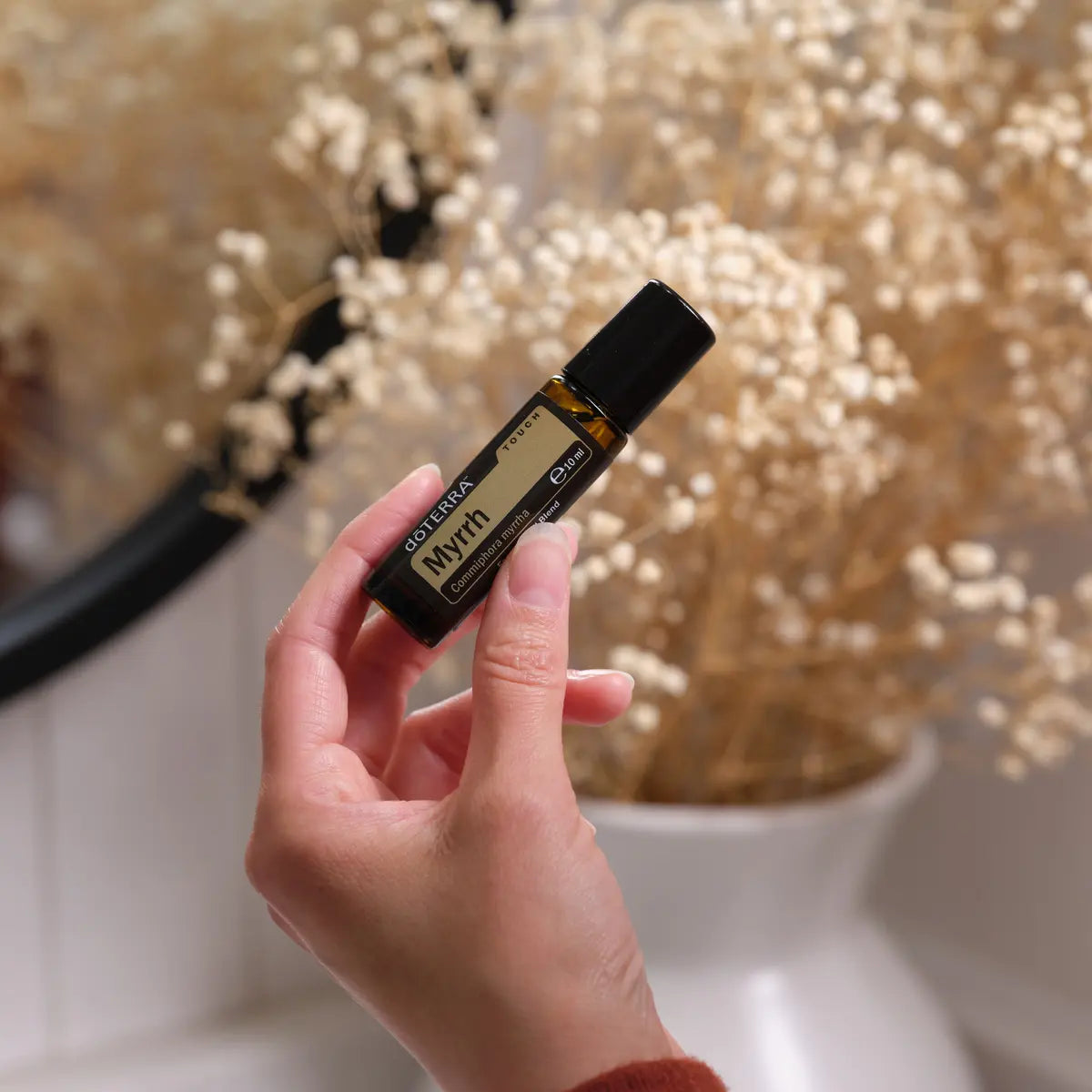 Mirra Touch dōTERRA roll-on 10ml en mano con flores secas gypsophila sobre fondo difuminado