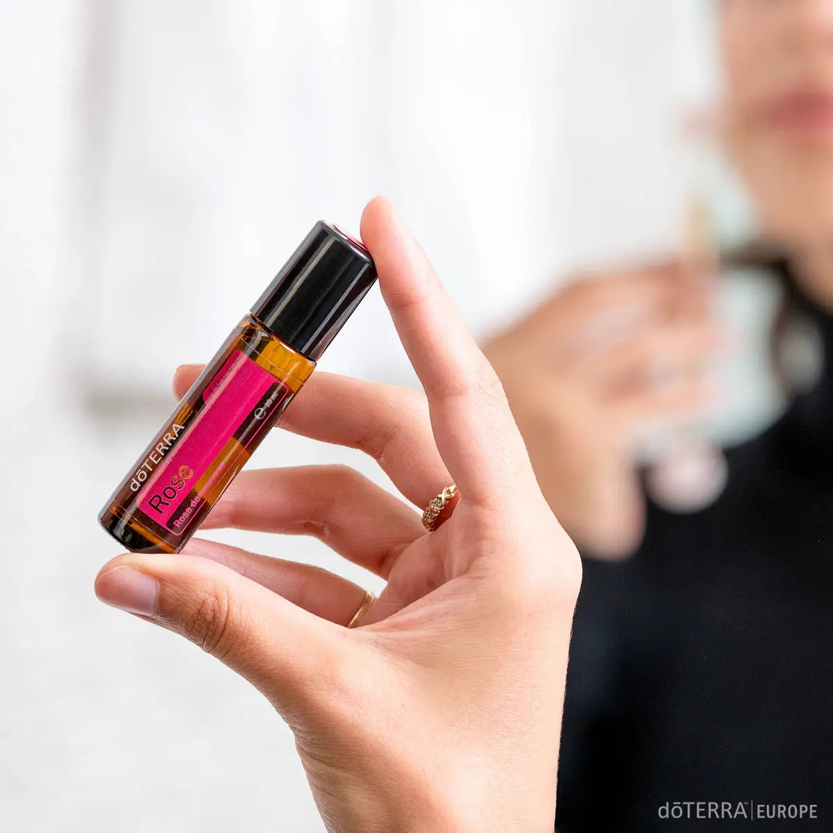 Rosa Touch dōTERRA roll-on 10ml sostenido por mano de mujer en interior con fondo bokeh