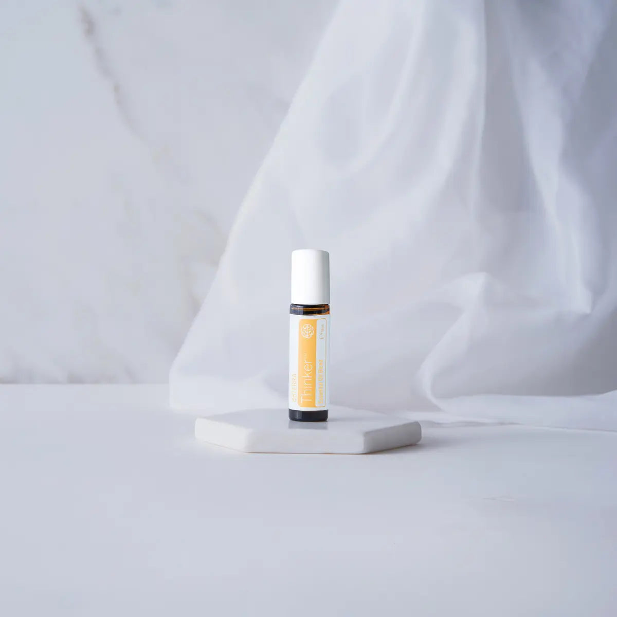 Roll-on Thinker dōTERRA 10ml — frasco sobre pedestal blanco con tela drapeada y mármol