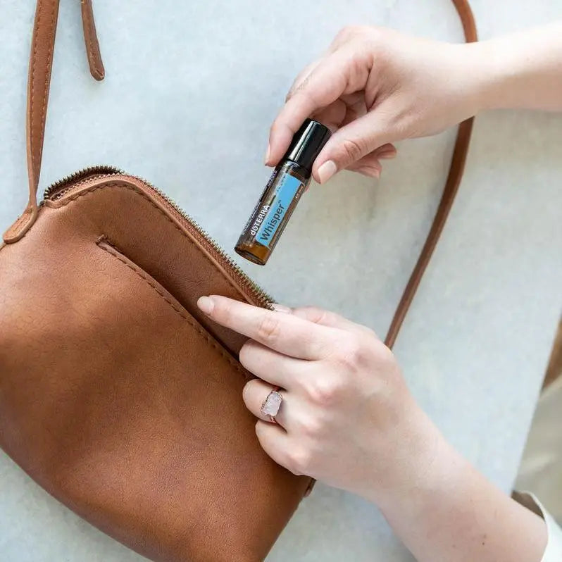 Roll-on Whisper Touch dōTERRA 10ml — manos sacando el frasco de un bolso de piel marrón