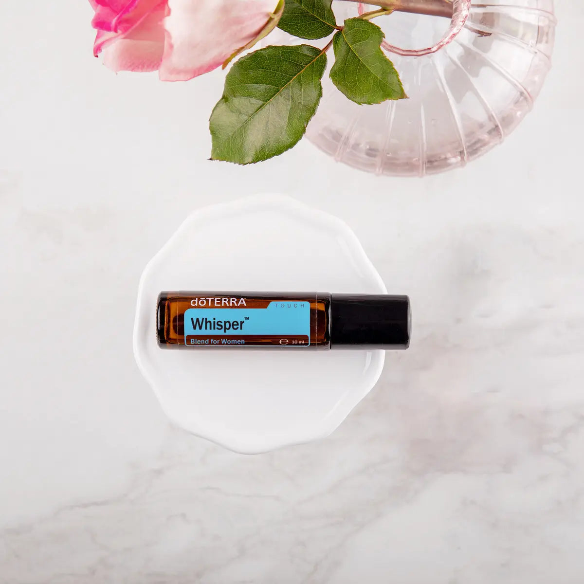 Roll-on Whisper Touch dōTERRA 10ml — frasco de cristal sobre plato ondulado rosa