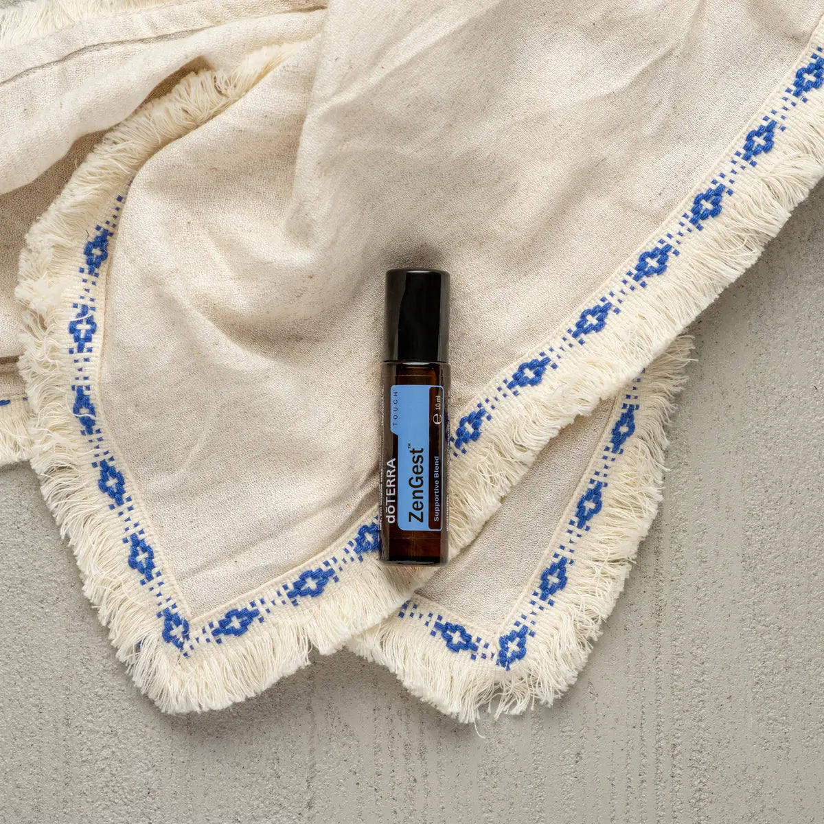 Roll-on ZenGest Touch dōTERRA 10ml — frasco sobre manta blanca con flecos y bordado azul