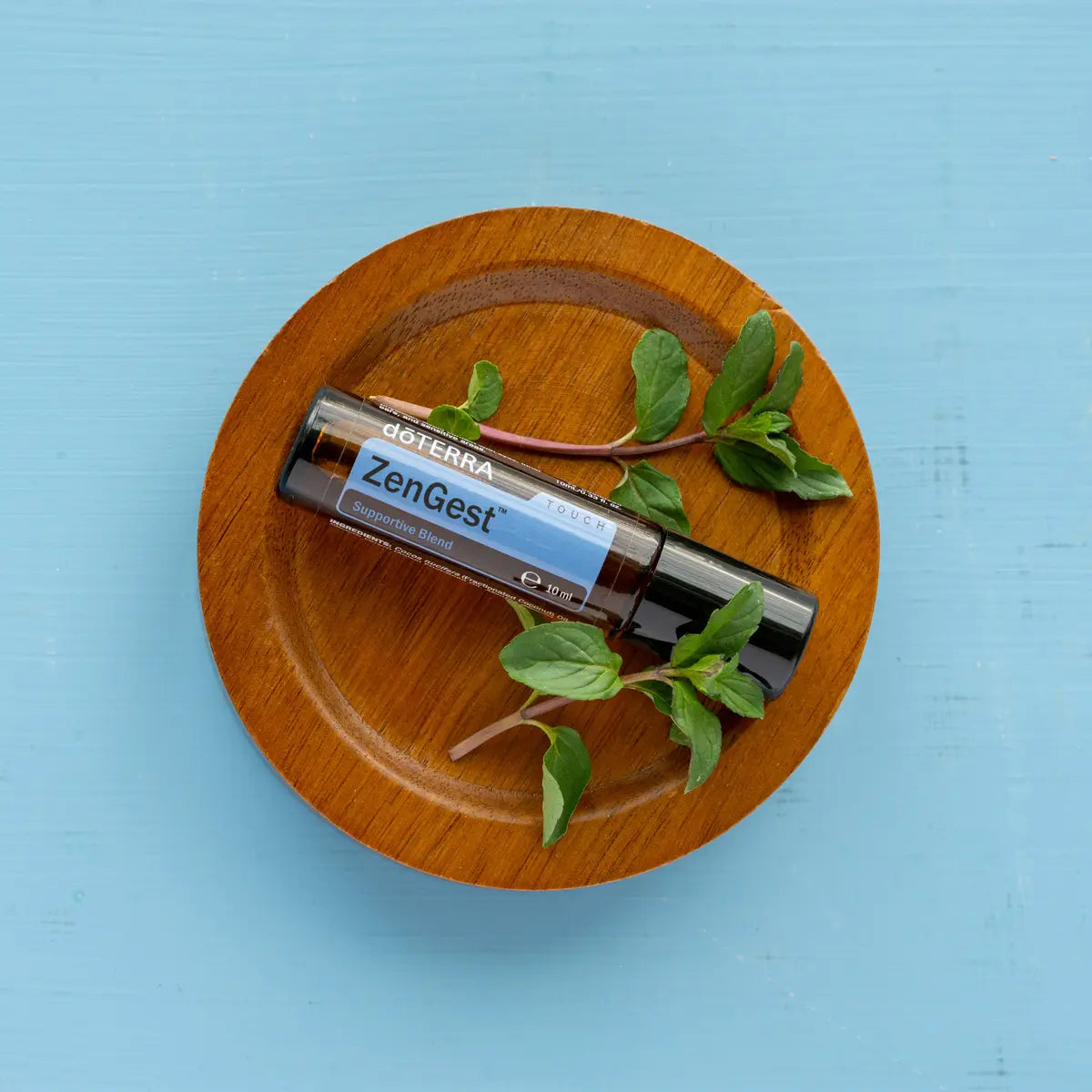 Roll-on ZenGest Touch dōTERRA 10ml — frasco sobre plato de madera con hierbabuena y fondo azul
