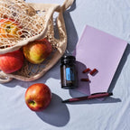 Frasco de Cápsulas Adaptiv dōTERRA junto a manzanas, bolsa y libreta rosa visto desde arriba