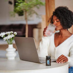 Mujer de cabello rizado bebiendo agua con frasco de Cápsulas Adaptiv dōTERRA en el escritorio