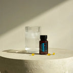 Frasco de DDR Prime Softgels dōTERRA junto a vaso de agua sobre pedestal de piedra con luz natural