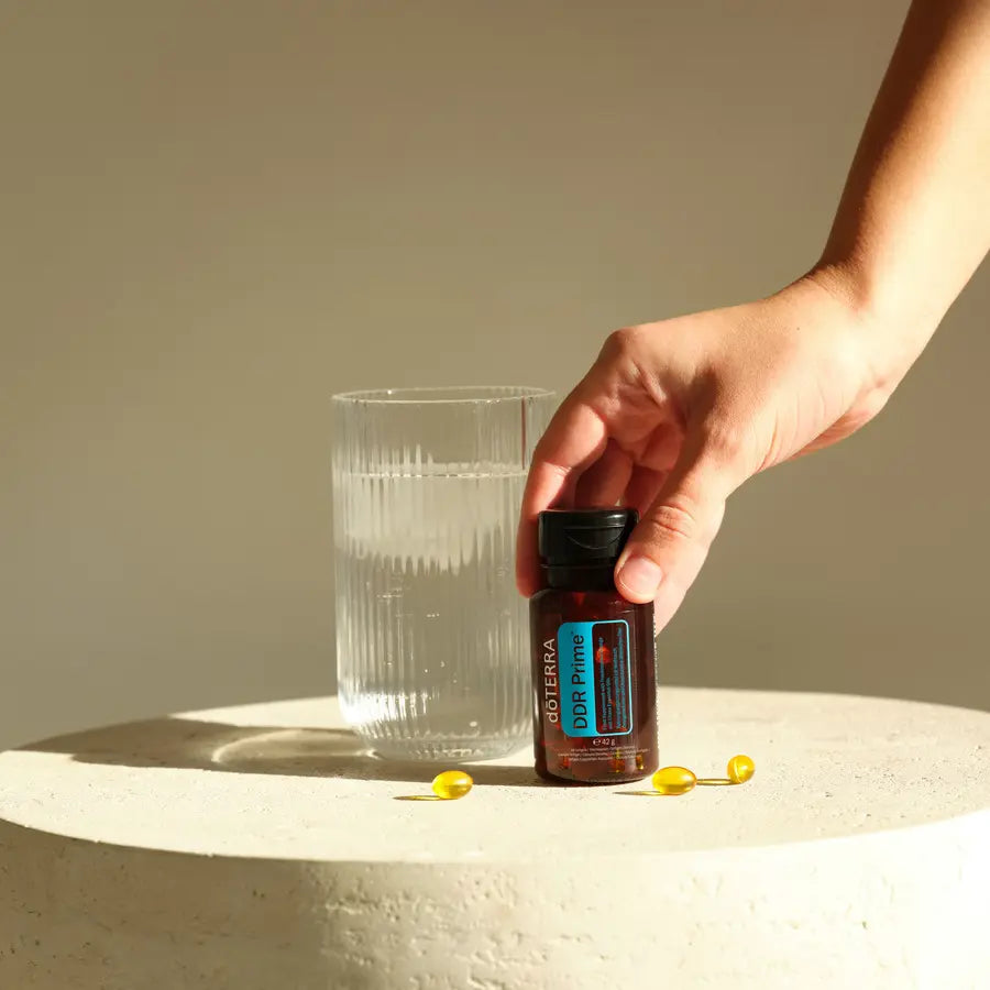 Mano cogiendo frasco de DDR Prime Softgels dōTERRA con vaso de agua sobre piedra