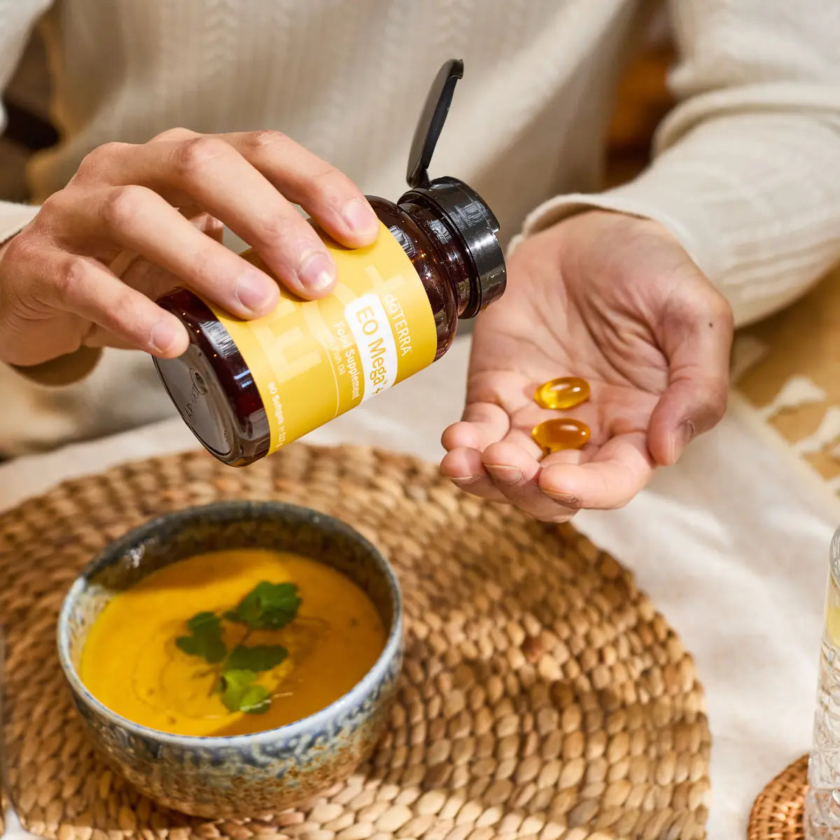 Manos sosteniendo frasco de Cápsulas EO Mega+ dōTERRA con softgels en cuenco de sopa