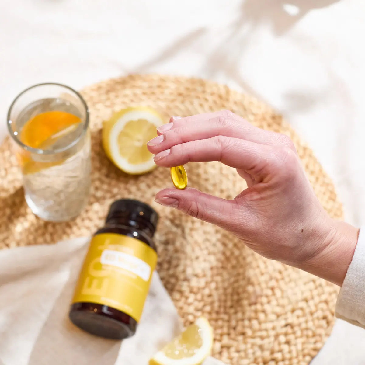 Mano con softgel de Cápsulas EO Mega+ dōTERRA junto a limón y vaso de agua en bandeja de mimbre